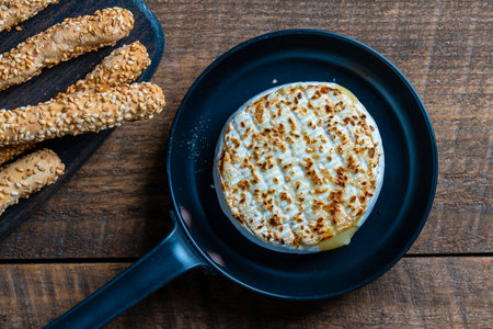 View of baked on frying pan camembert cheese and bread sticks with sesame seeds on wooden table background, close up, top viewの写真素材