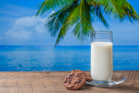 Fresh white milk in a glass with cookies on a wooden table with sea water, coconut palm tree and blue sky background on sunny summer day in tropical beach cafe, close upの写真素材