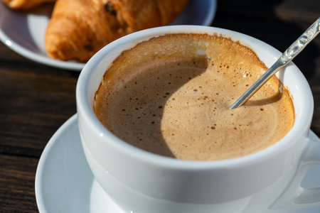 Thick foam of hot coffee cappuccino with spoon in white cup and croissant on wooden table, close up....