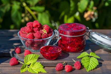 Red raspberry jam and fresh raspberry on a rustic wooden table outdoors near garden. Rustic style,...