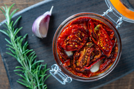 Sun-dried red tomatoes with garlic, green rosemary, olive oil and spices in a glass jar on a wooden table. Rustic style, top view, close upの写真素材