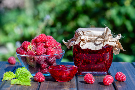 Red raspberry jam and fresh raspberry on a rustic wooden table outdoors near garden. Rustic style,...