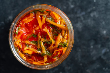 Delicious pickled tomatoes with carrots, garlic and parsley in a glass jar on a dark background, close up, top view, copy spaceの写真素材