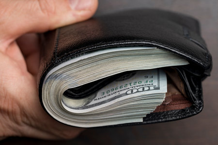 A man's hand holds a black leather wallet with a bunch of US 100 dollars bills, banknotes, cash...