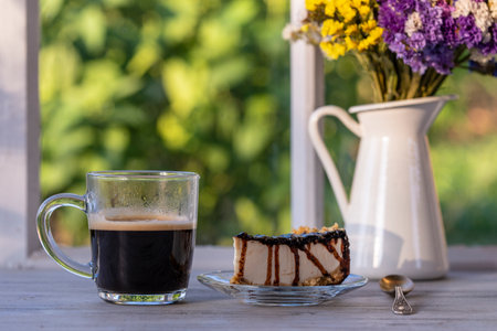 Cheesecake with chocolate sauce and glass cup of black coffee on a wooden table near window, close up. Coffee time with slice of cake at morningの写真素材