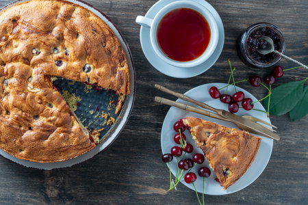 Cut piece of cherry sponge pie along with fresh cherries and white cup of tea on a wooden table, top view, close up. Delicious summer homemade American cake with cherries. Sweet dessert for breakfastの写真素材
