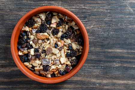 Whole grain oat muesli with dried fruits, nuts, raisins and berries in clay bowl on wooden table, top view, close up, copy space. Background of organic muesli for breakfastの写真素材