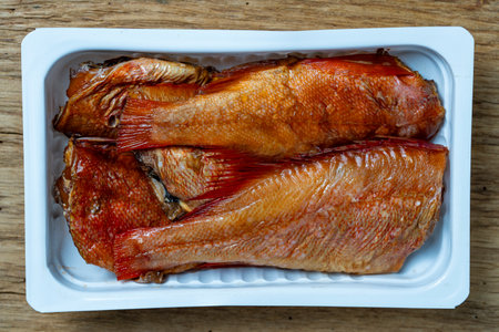 Hot smoked perch fish in white plastic packaging on wooden background. Close up, top view