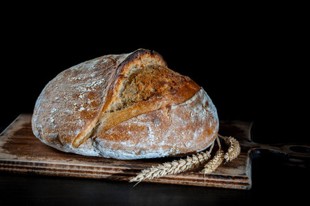 Round loaf of freshly baked sourdough bread with ears of wheat on wooden cutting board, close up....