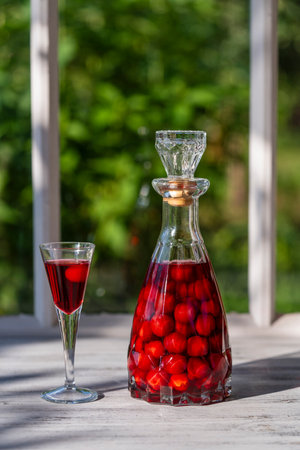 Homemade cherry brandy in wine glass and in a glass bottle on a wooden windowsill near summer garden, close upの写真素材