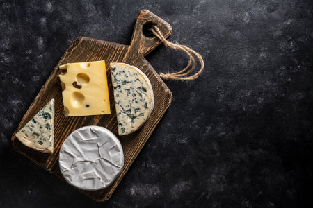 Set of different cheeses on wooden cutting board on a black background, close up, top view, copy...