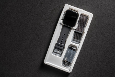 Black smart watch with a belt in a white plastic package on black table, close up, top viewの写真素材