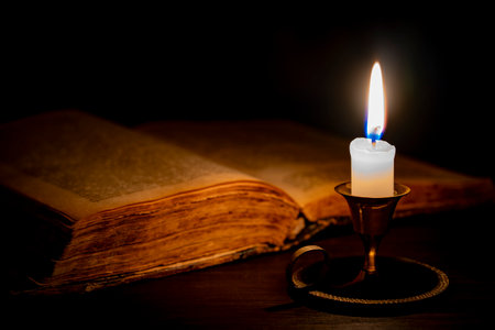 Burning wax candle in bronze candlestick on wooden table next to old open book in dark room at night, close up. Vintage still life with lighting candle near old bookの写真素材