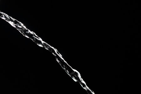 Crystal clear stream of water flowing on isolated black background, close up. Transparent pouring stream of clean water cascading against a dark backdrop. Concept of drinking pure waterの写真素材