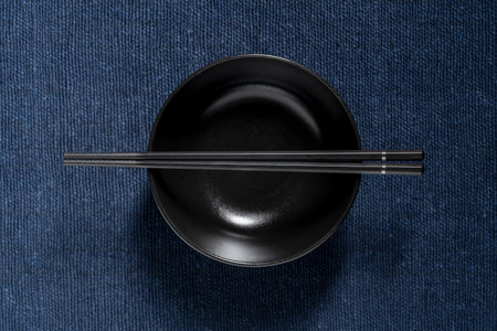 A pair of chopsticks lying across an empty black soup bowl on blue fabric background, close up, top viewの写真素材