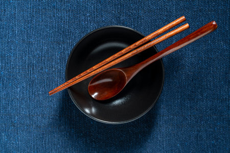 A pair of wooden chopsticks and spoon lying across an empty black soup bowl on blue fabric background, close up, top viewの写真素材