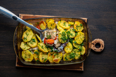 Sliced baked potato, fish fillet pollock, tomatoes, onion and sprinkled herbs in a glass baking dish on a wooden table, close up, top viewの写真素材