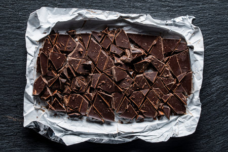 Broken dark chocolate bar on wrapping foil on black background, close up, top view. Sweet food