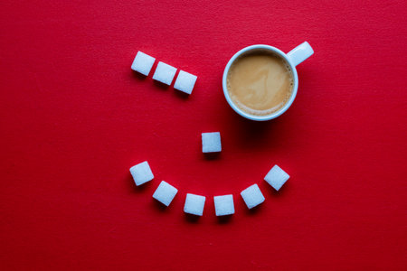 Happy face shape made from white cup of espresso coffee and sugar cubes on red background, close up,...
