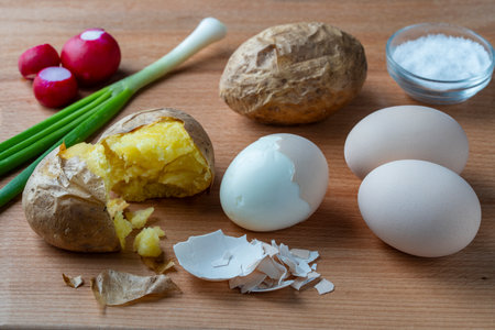 Broken baked potatoes, hard boiled eggs, green onions and radishes on a wooden table, close up....