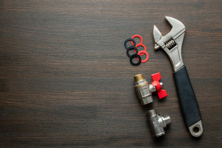 Metal adjustable wrench, ball valve with red handle, stainless steel tee fittings and rubber gaskets on wooden background, close up, top view, copy space. Plumbing repair conceptの写真素材