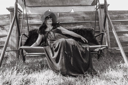 Portrait of a happy elderly woman 65 - 70 years old in a straw hat resting on a swing in the garden,...