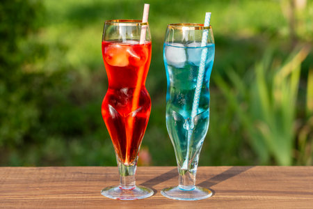 Two artistic glasses shaped like a nude woman and man filled with red and blue alcoholic cocktails on table with lush green garden background, close up. Playful summer vibe for creative drink conceptsの写真素材