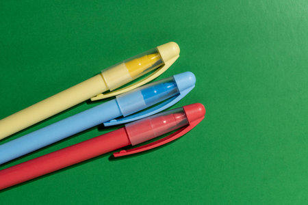 Close up of yellow blue and red plastic pens lying diagonally on bright green surface. Minimalist office stationery and back to school concept with copy space. Three colorful pens on green background