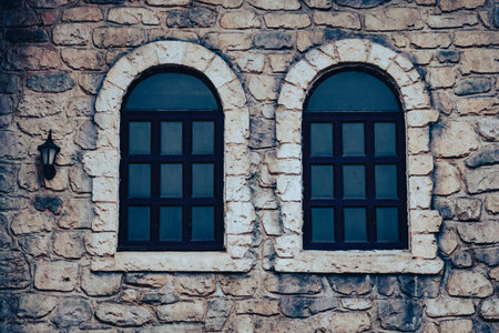 Two arched vintage windows on a rustic stone wall of an old house. Symmetrical shot of two classic arched windows with dark frames built into an ancient rough stone facade with a small lantern