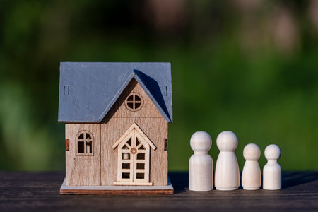 Small miniature house model with a grey roof stands beside a wooden peg doll family on desk with blurred green nature background representing real estate and safety, close up, outdoors