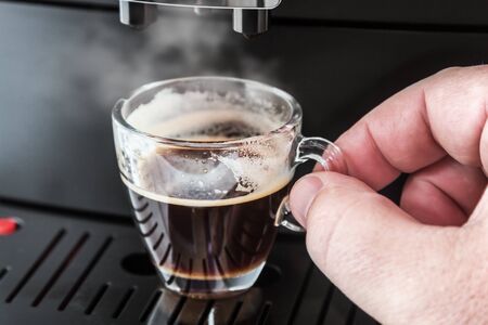 freshly brewed coffee is poured from the coffee machine into glass cup. Hand holding Cupの写真素材