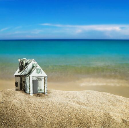 house carved out of dollar bills on the sea sand. Concept on the topic of selling or buying a house, buying a house on the sea or ocean shoreの写真素材