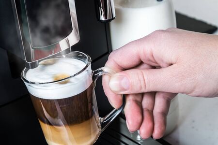 hand picks up a glass of hot cappuccino with foam made in the coffee machineの写真素材