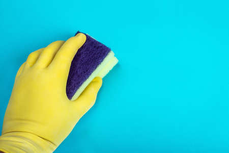 hand in a yellow rubber glove holds a sponge for washing and cleaning on a blue background