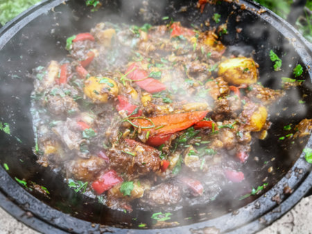 Delicious spicy beef stew cooking in a traditional pot outdoors with fresh vegetables and herbsの写真素材