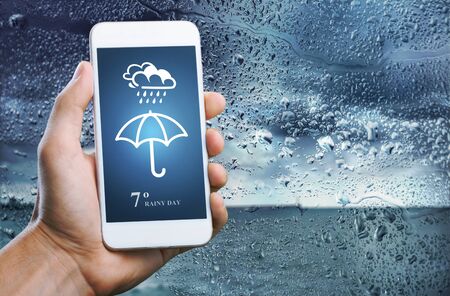 Rainy Day Concept. Hand Holding Smartphone with Weather Information show on Screen. Blurred Traffic Jam and Rain Drops on Glass Window as backgroundの写真素材