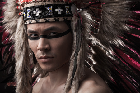 Naked indian strong man with traditional native american make up and headdress looking at the camera. Close up desaturated studio shotの写真素材