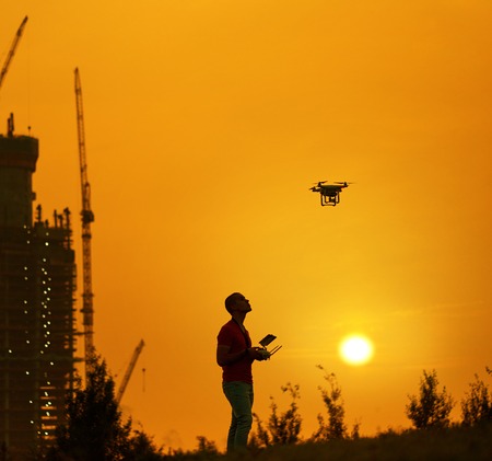 Man controls a quadrocopter at the warm sunset. New building on background.