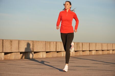 A jogging woman in red running shirt and black leggins on the street at sunrise. Running on concrete quayの写真素材