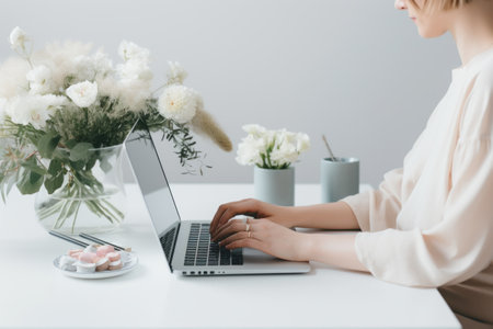 a woman's hands on her laptop as she looks at some flowers on a desk