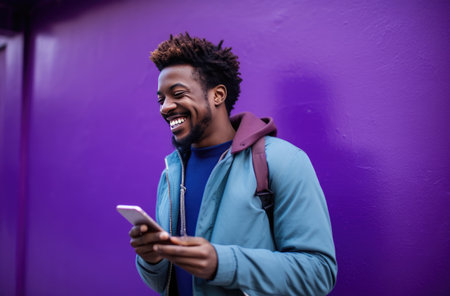 a smiling man looking at his phone while standing on a purple street wall.の素材