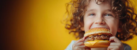 A young girl sitting and eating a hamburger against a vibrant yellow backdrop.の素材