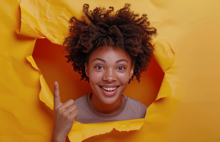 A woman with curly hair smiles broadly as she peeks through a torn hole in a yellow paper backdrop. She is pointing with her right index finger.の素材