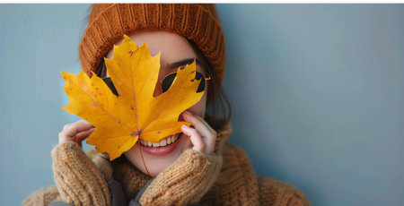A woman in a brown coat holds a yellow maple leaf in front of her face, against a light blue wall.の素材