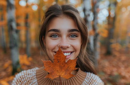 A woman smiles happily while holding a bright orange leaf in front of her face in a forest setting.の素材