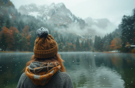 A woman wearing a knit hat and scarf looks out at a mountain lake as snow falls around her.の素材