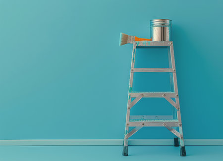 A silver ladder stands against a light blue wall with three blue paint cans and a paintbrush resting on it.の素材