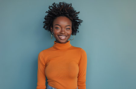 A woman with curly hair smiles while wearing an orange turtleneck sweater against a light blue backdrop.の素材