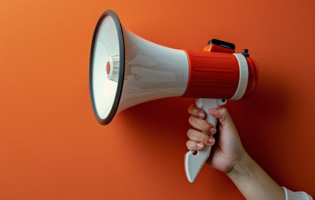 A person wearing a hooded jacket holds an orange megaphone in front of a gray wall.の素材