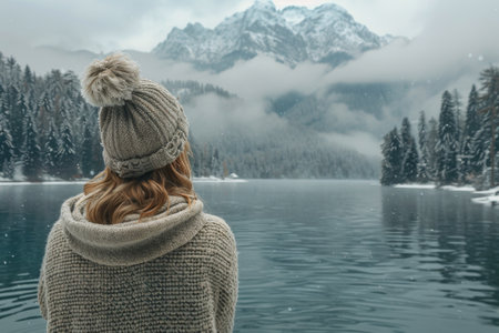 A woman wearing a knit hat and sweater stands looking at a snowy mountain lake, with snow falling and fog in the distance.の素材